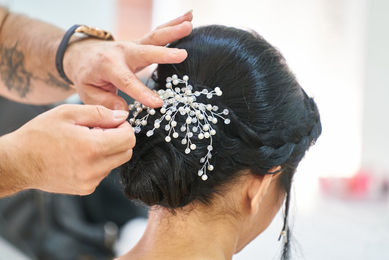 hero-img-02 Photograph of a hairstylist arranging an elegant updo embellished with delicate decorative pins in a professional studio.