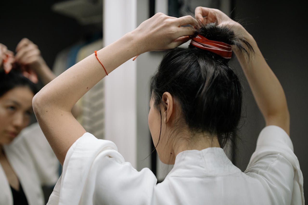 gallery-2 Woman in bathrobe styling hair in front of mirror, focusing on hair bun.